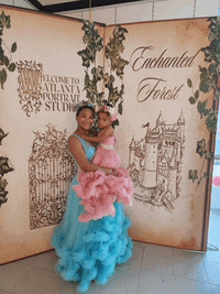 a woman in a blue and pink dress holds a little girl in front of a book that says enchanted forest