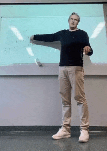 a man in a black sweater and khaki pants is standing in front of a whiteboard .