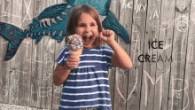 a little girl is holding a cone of ice cream in front of a wooden fence with the word ice cream written on it