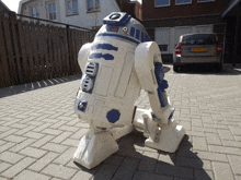 a r2d2 statue sits on a brick driveway next to a silver car