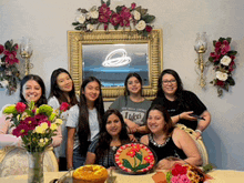 a group of women standing around a table with flowers and a cake