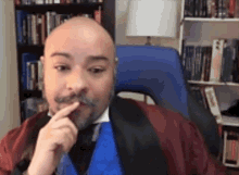 a man with a beard and mustache is sitting in front of a bookshelf and looking at the camera .