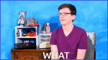 a man wearing glasses and a purple shirt is sitting in front of a desk with the words what on it