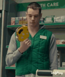 a man in a green vest is holding a yellow object in front of a shelf that says health care .