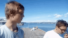 two boys are standing on a dock near the water and one of them is smoking a cigarette