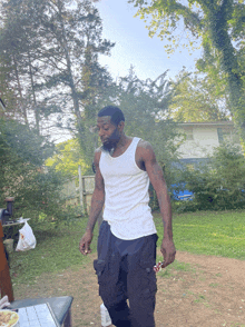 a man in a white tank top and black pants is standing in a yard