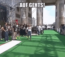 a group of people are standing on a green carpet with the words auf gehts written on the bottom .