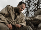 a man in a tan jacket is sitting on a pile of rocks looking down .