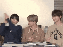 three young men are sitting at a table with one wearing a bathing ape shirt