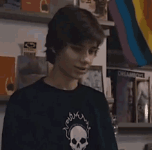 a young man wearing a black shirt with a skull on it is standing in front of a shelf with books .