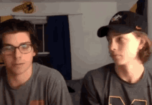 two young men are sitting next to each other in a living room . one of the men is wearing glasses and a hat .