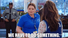 a woman in a blue shirt is talking to another woman in a store while a security guard stands behind them .