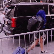 a woman with blue hair is pushing a blue suitcase behind a fence with the next thing written on it