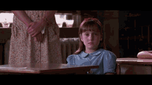 a little girl in a blue shirt sits at a desk in front of a woman in a floral dress