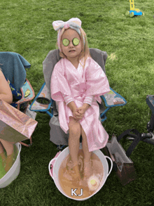 a little girl in a pink robe with cucumber slices on her eyes is sitting in a chair