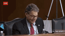 a man in a suit and tie sits at a table with a name plate that says robert franken on it