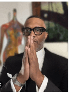 a man wearing glasses and a suit is praying with his hands folded .