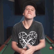 a young man wearing a black shirt with a heart on it is sitting in front of a pool table .