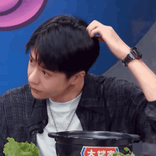 a young man adjusts his hair while sitting in front of a pot of food