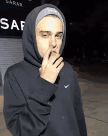 a man in a hoodie is smoking a cigarette while standing on a sidewalk .