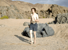 a shirtless man in swim trunks walking on a beach