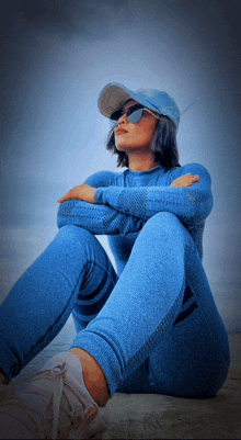 a woman wearing a blue hat and sunglasses sits on the ground with her arms crossed