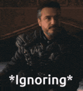 a man in a leather jacket is sitting on a bed with the words * ignoring * written below him .