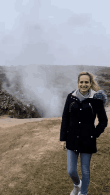 a woman in a black coat is standing in front of a mountain with smoke coming out of it