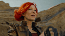 a woman with red hair and a brown jacket stands in the desert