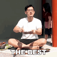 a man sits in a lotus position holding a bowl of food with the words " the best " behind him