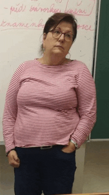 a woman in a pink striped shirt and glasses is standing in front of a white board .