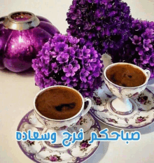 two cups of coffee on saucers next to purple flowers and a purple vase