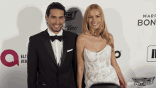 a man in a tuxedo and a woman in a white dress pose for a photo on a red carpet that says make bon