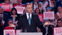 a man in a suit and tie stands at a podium with a crowd behind him holding signs that say duda 2020