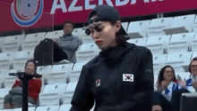 a woman wearing sunglasses and a baseball cap is standing in a stadium with a flag on her shirt .