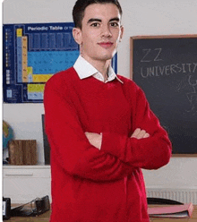 a young man in a red sweater is standing in front of a blackboard with his arms crossed in a classroom .