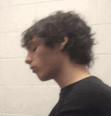 a young man with curly hair and a black shirt is standing in front of a white wall .