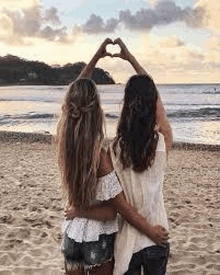 two women are standing on a beach making a heart shape with their hands .