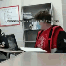 a boy in a red shirt is sitting at a desk in a classroom with a laptop and a book .