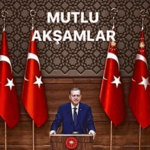 a man in a suit and tie is standing at a podium with flags behind him and the words mutlu aksamlar above him