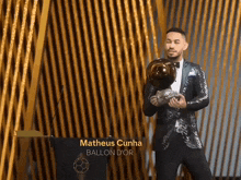 a man in a suit holds a soccer ball in front of a podium with matheus cunha ballon d' or written on it