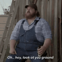 a man with a beard wearing overalls and a plaid shirt is sitting on a ladder holding a beer .