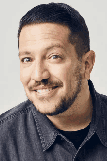 a man with a beard wearing a denim shirt smiles