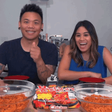 a man and a woman sit at a table with bowls of food and a bag of hot chicken noodles