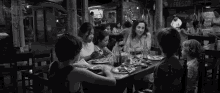 a black and white photo of a group of people sitting at a table in a restaurant eating food .