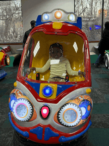 a little girl sits in a colorful toy car with a red light on the front