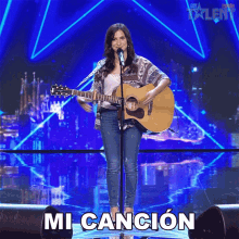 a woman singing into a microphone while holding a guitar in front of a sign that says ' mi cancion '