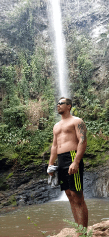 a shirtless man stands in front of a waterfall wearing adidas shorts