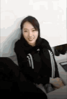 a woman in a black hoodie is smiling while sitting on a couch .