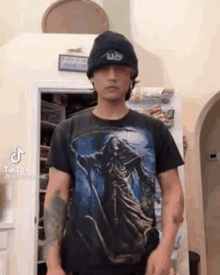 a young man wearing a black hat and a black t-shirt is standing in a kitchen .